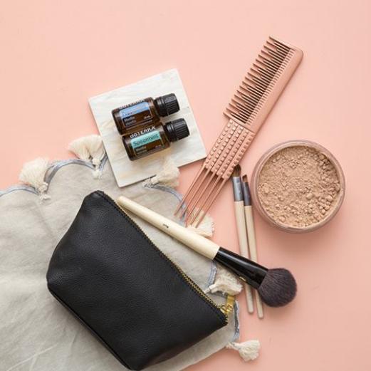 Neceser de maquillaje negro con brochas, peine profesional y aceites esenciales dōTERRA sobre un fondo rosa suave.