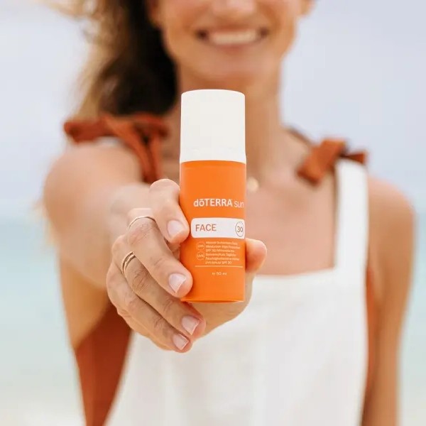 Persona sosteniendo el protector solar dōTERRA sun Face SPF 30 en la playa con fondo desenfocado.