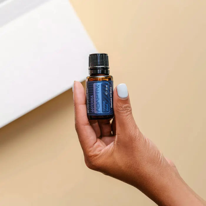 Mano sosteniendo un frasco de aceite esencial dōTERRA Adaptiv de 15ml sobre un fondo minimalista de tonos neutros.