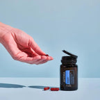 Mano sosteniendo una cápsula de Adaptiv™ dōTERRA junto al frasco abierto, con cápsulas de color ámbar sobre un fondo azul claro minimalista; suplemento para la calma y la gestión del estrés.