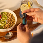 Manos sosteniendo un frasco de 15 ml de dōTERRA RevitaZen junto a un tazón de yogurt con granola, kiwi y un vaso de agua con limón.