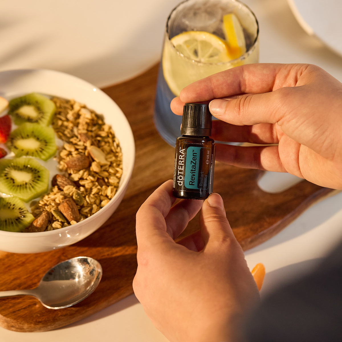 Manos sosteniendo un frasco de 15 ml de dōTERRA RevitaZen junto a un tazón de yogurt con granola, kiwi y un vaso de agua con limón.