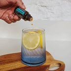 Mano virtiendo una gota del aceite esencial dōTERRA RevitaZen en un vaso con agua fresca y rodajas de limón sobre una tabla de madera.