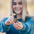 Mujer rubia sonriendo sosteniendo frasco de aceite esencial de Abeto Siberiano dōTERRA 15ml con ambas manos, jersey azul, exterior desenfocado