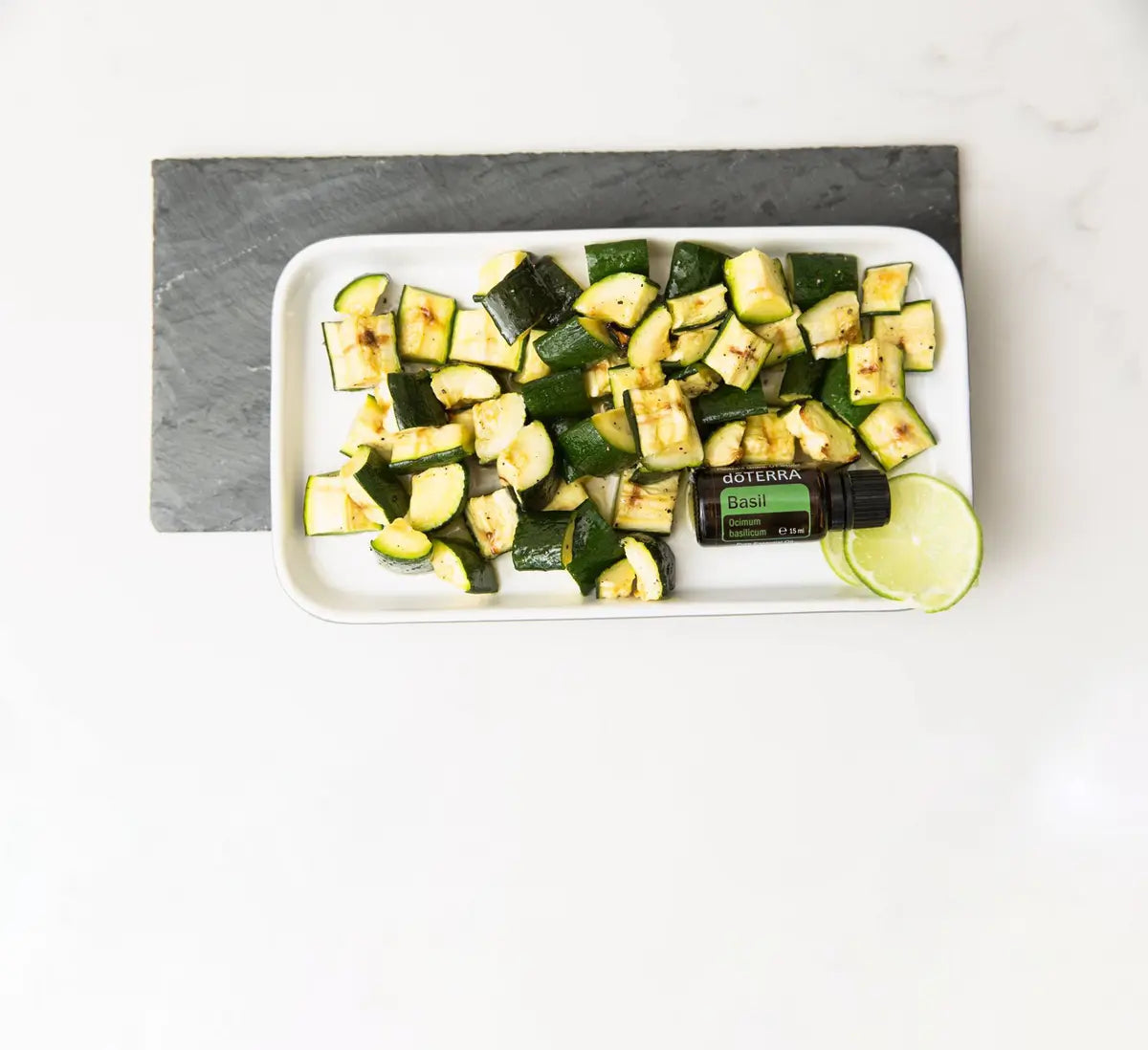 Vista aérea del Aceite Esencial de Albahaca dōTERRA 15ml sobre plato blanco con calabacín a la plancha y rodaja de lima, base de pizarra gris