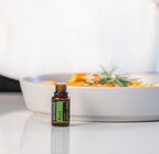 Frasco del Aceite Esencial de Albahaca dōTERRA 15ml sobre superficie blanca reflectante, cazuela con pasta y romero desenfocados al fondo