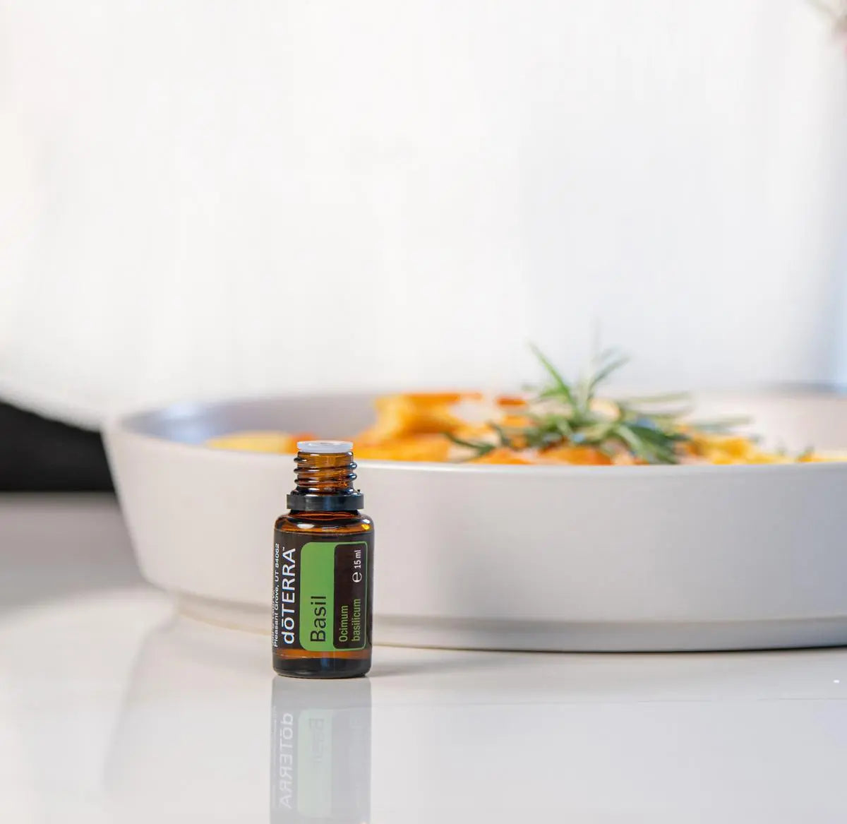 Frasco del Aceite Esencial de Albahaca dōTERRA 15ml sobre superficie blanca reflectante, cazuela con pasta y romero desenfocados al fondo