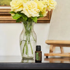 Frasco del Aceite Esencial de Árbol de Té dōTERRA 15ml sobre consola negra junto a florero de vidrio acanalado con rosas amarillas y caballete de madera, cuadro al fondo