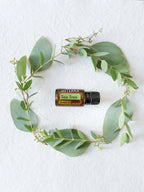 Vista cenital del Aceite Esencial de Árbol de Té dōTERRA 15ml en el centro de una corona de ramas de eucalipto con hojas redondeadas, sobre tela blanca texturada
