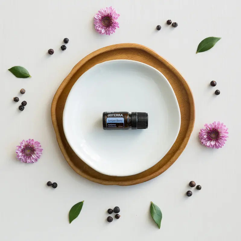 Aceite esencial Juniper Berry dōTERRA 5ml en plato blanco sobre bandeja de madera con bayas de enebro y flores lilas, vista cenital