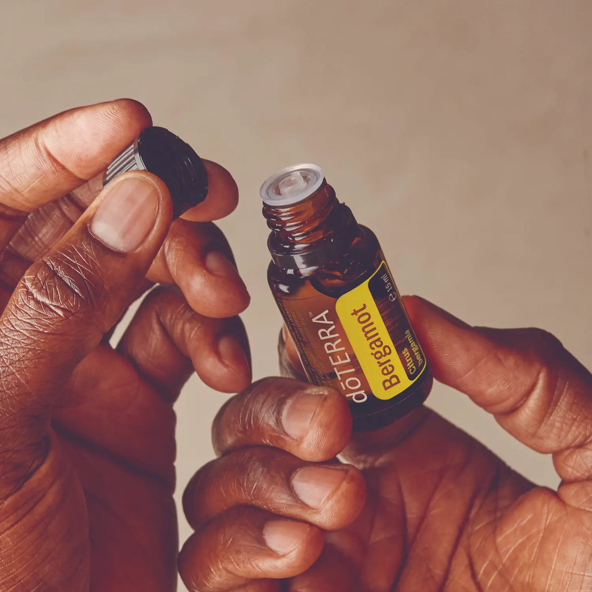 Manos de piel oscura abriendo el frasco del Aceite Esencial de Bergamota dōTERRA 15ml con la tapa retirada, fondo beige cálido
