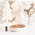 Frasco del Aceite Esencial de Canela dōTERRA 5ml junto a cuenco blanco con granola y arándanos secos, árboles decorativos de lana y jarrón de algodón en flor al fondo