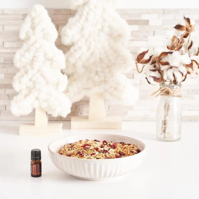 Frasco del Aceite Esencial de Canela dōTERRA 5ml junto a cuenco blanco con granola y arándanos secos, árboles decorativos de lana y jarrón de algodón en flor al fondo