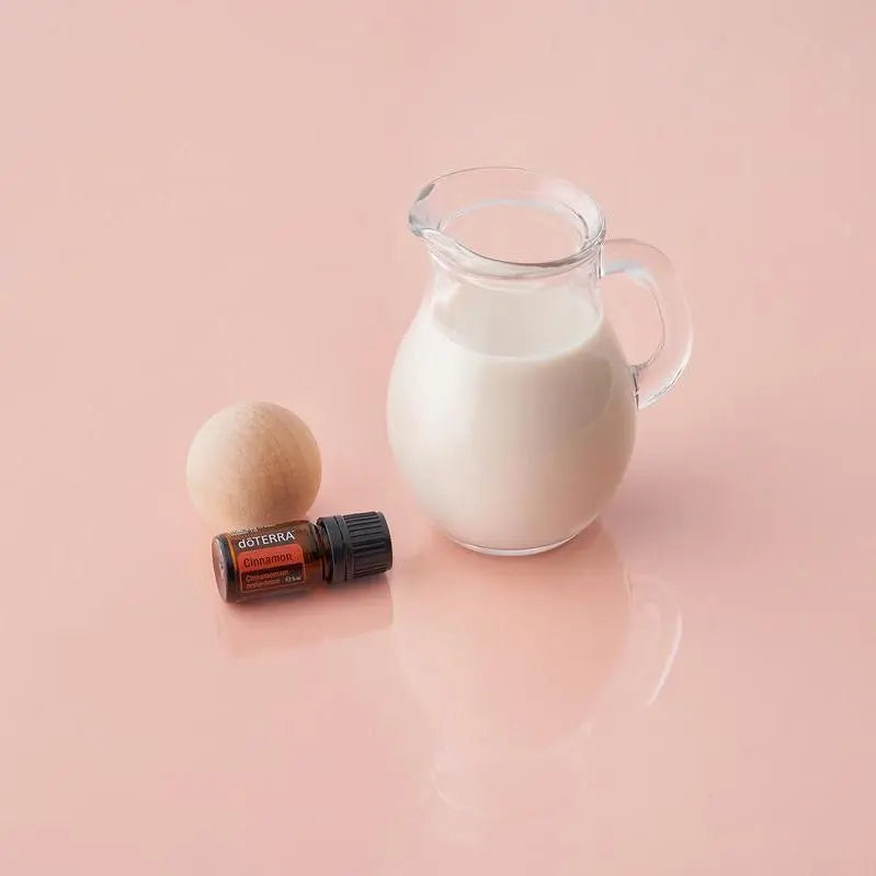 Frasco del Aceite Esencial de Canela dōTERRA 5ml con bola de madera junto a jarra de vidrio con leche, superficie y fondo rosa rosado
