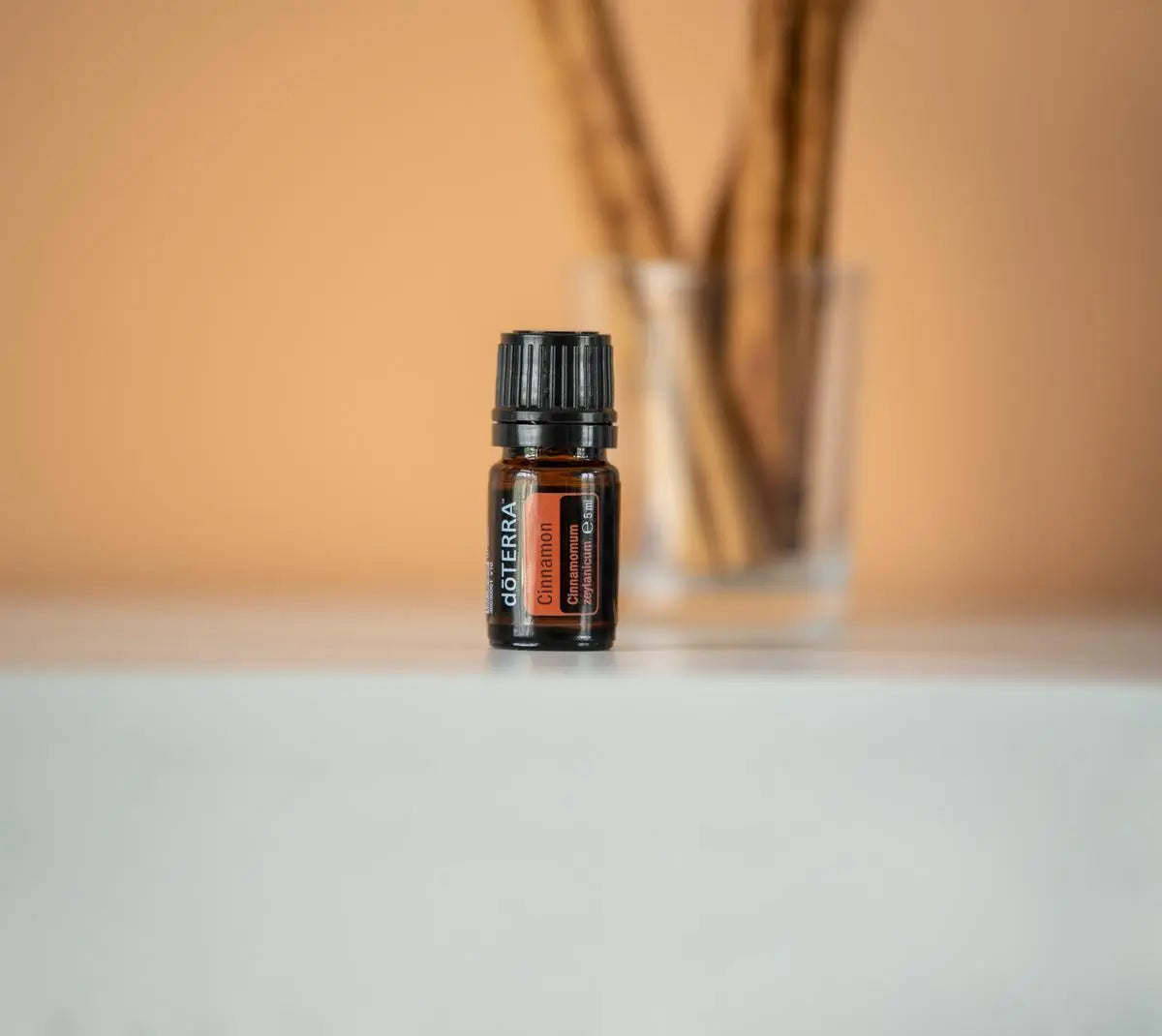 Frasco del Aceite Esencial de Canela dōTERRA 5ml sobre superficie blanca con difusor de mimbres en vaso de vidrio desenfocado al fondo, tono beige cálido