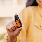 Mujer con chaqueta mostaza sosteniendo el Aceite Esencial de Casia dōTERRA 15ml hacia la cámara, fondo exterior desenfocado