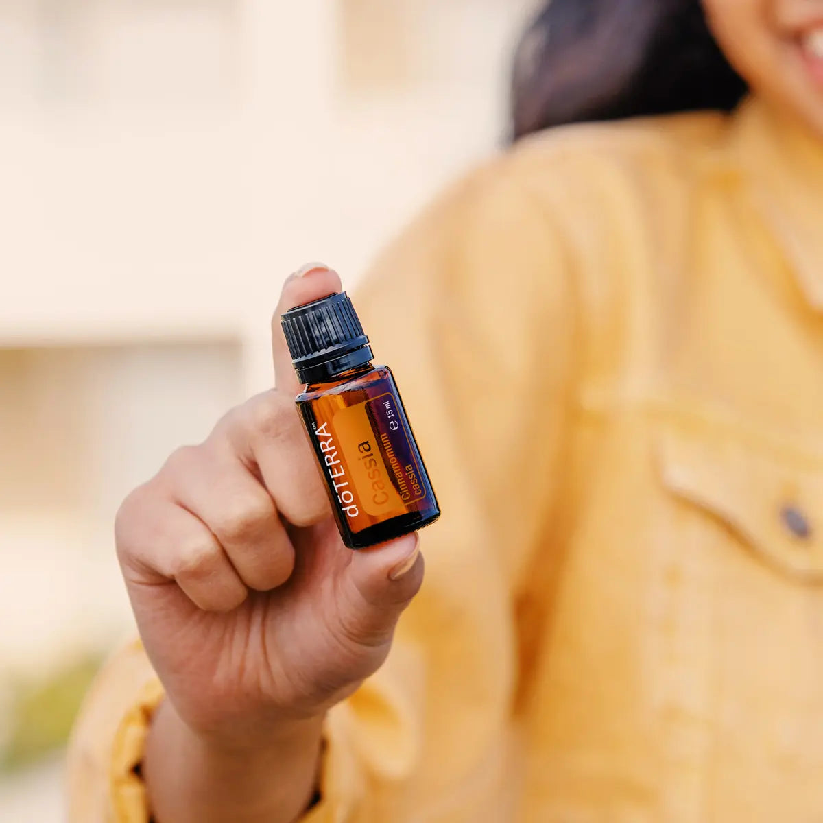 Mujer con chaqueta mostaza sosteniendo el Aceite Esencial de Casia dōTERRA 15ml hacia la cámara, fondo exterior desenfocado