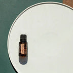 Frasco del Aceite Esencial de Cedro dōTERRA 15ml sobre bandeja redonda blanca con fondo verde oscuro y luz solar lateral