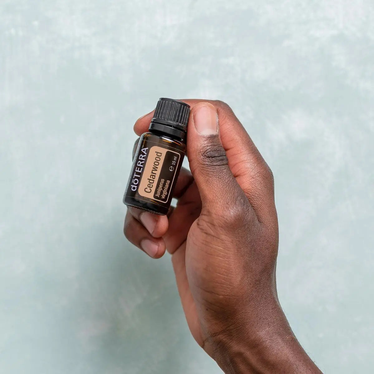 Mano de piel oscura sujetando el Aceite Esencial de Cedro dōTERRA 15ml ante pared de textura gris azulada