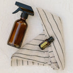 Frasco del Aceite Esencial de Citronela dōTERRA 15ml sobre tela de lino a rayas junto a spray de vidrio ámbar, pared blanca texturada al fondo