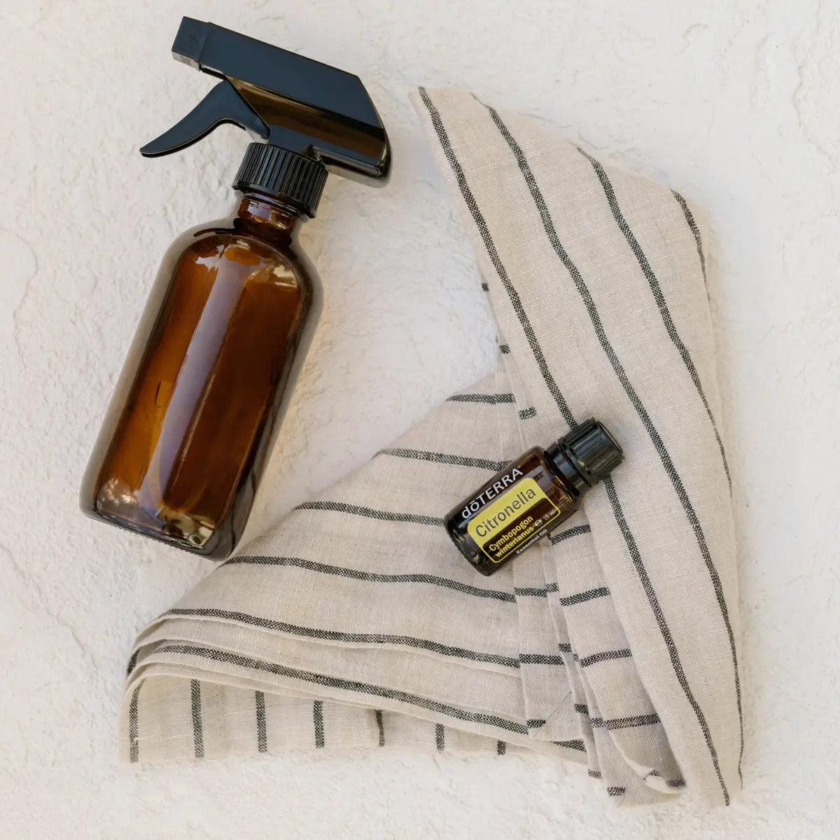 Frasco del Aceite Esencial de Citronela dōTERRA 15ml sobre tela de lino a rayas junto a spray de vidrio ámbar, pared blanca texturada al fondo