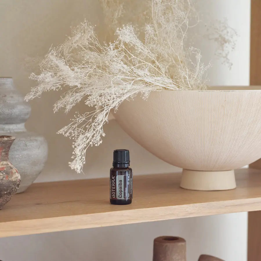 Frasco del Aceite Esencial de Copaiba dōTERRA 15ml en estante de madera clara junto a cuenco cerámico estriado beige con flores de gypsophila secas blancas y jarras de barro antiguas