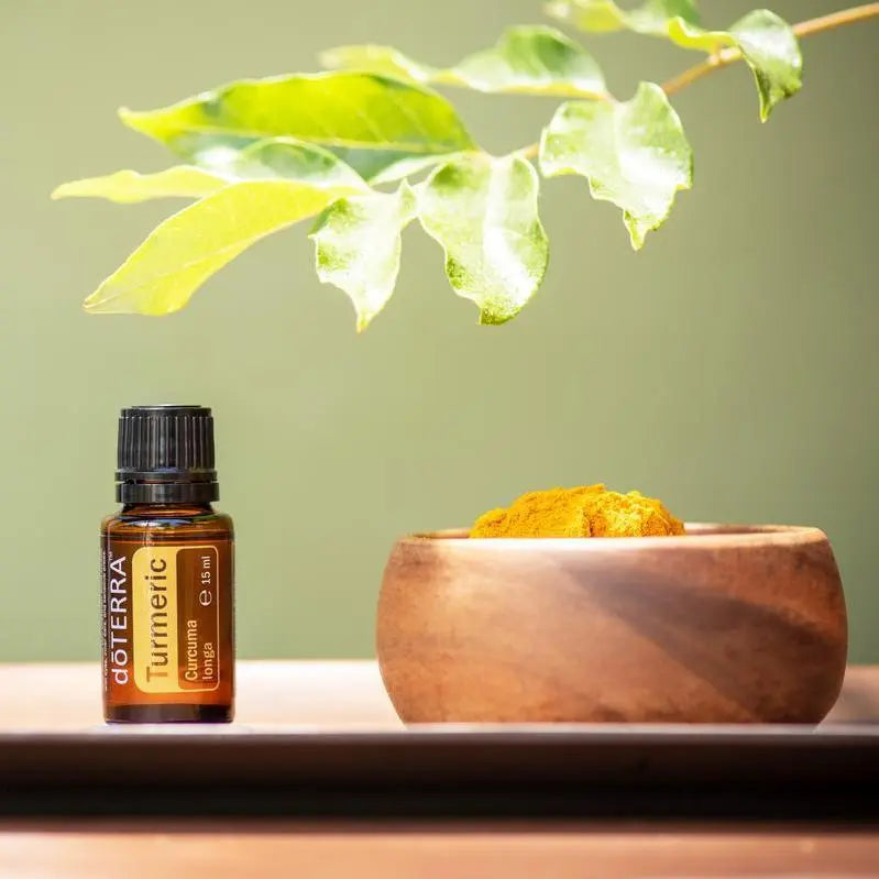 Frasco del Aceite Esencial de Cúrcuma dōTERRA 15ml en bandeja oscura junto a cuenco de madera con polvo de cúrcuma, hoja verde variada colgando desde arriba, fondo verde salvia