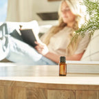 Frasco del Aceite Esencial de Cúrcuma dōTERRA 15ml en primer plano sobre mesa de madera de salón, mujer rubia leyendo un libro en sofá blanco desenfocada al fondo con rama de ciprés