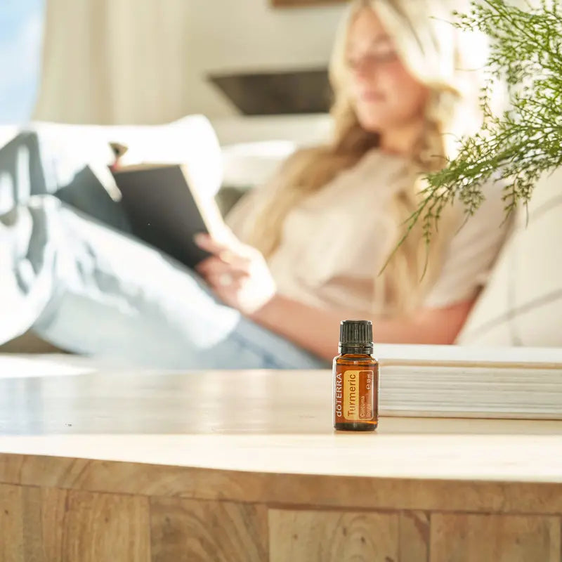 Frasco del Aceite Esencial de Cúrcuma dōTERRA 15ml en primer plano sobre mesa de madera de salón, mujer rubia leyendo un libro en sofá blanco desenfocada al fondo con rama de ciprés