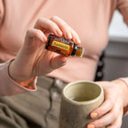 Manos femeninas con uñas melocotón inclinando el Aceite Esencial de Cúrcuma dōTERRA 15ml sobre taza de cerámica rústica, jersey rosa al fondo