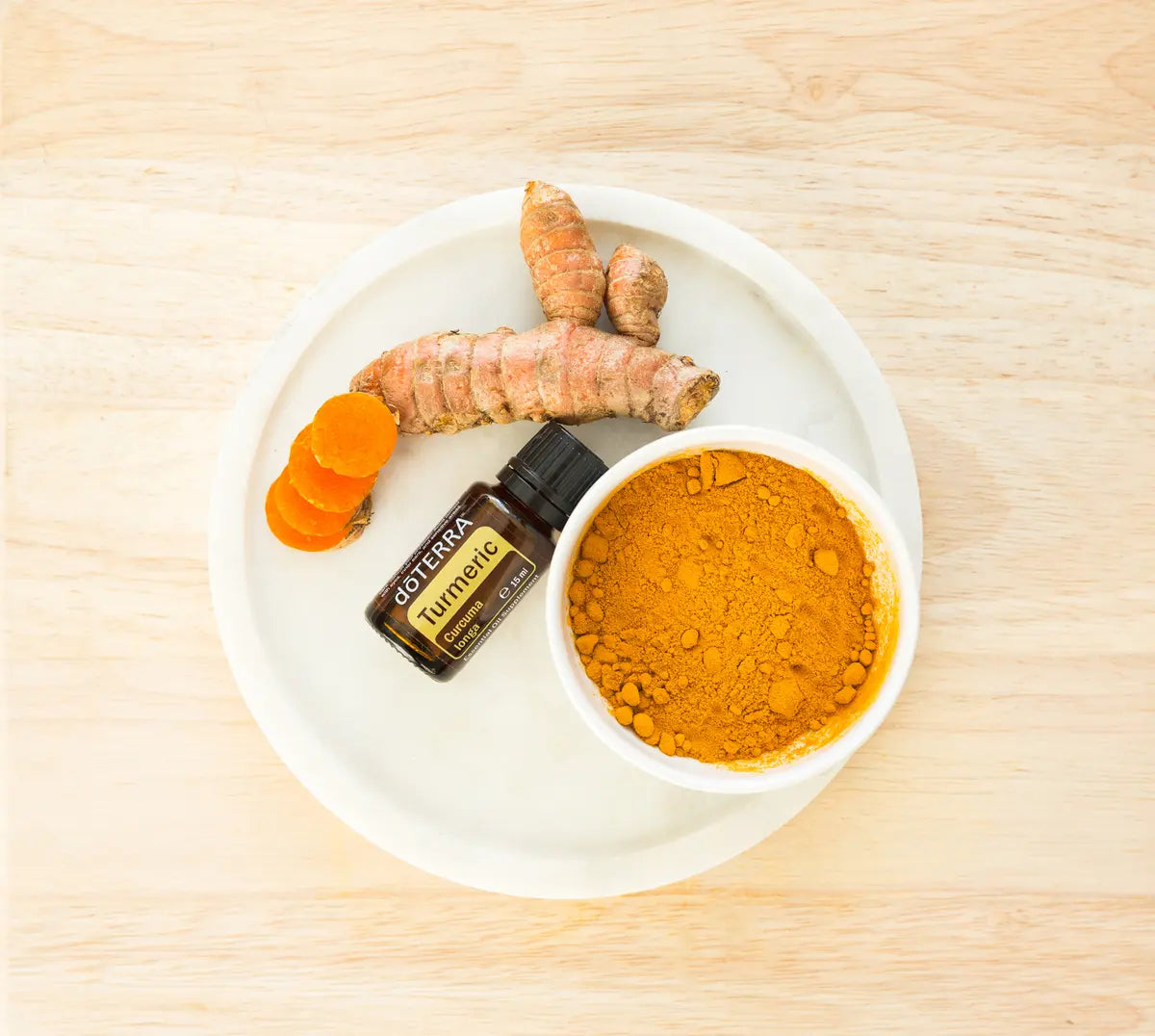 Vista cenital del Aceite Esencial de Cúrcuma dōTERRA 15ml en plato de mármol blanco junto a raíz de cúrcuma entera, rodajas naranjas y cuenco con polvo de cúrcuma dorado, superficie de madera clara