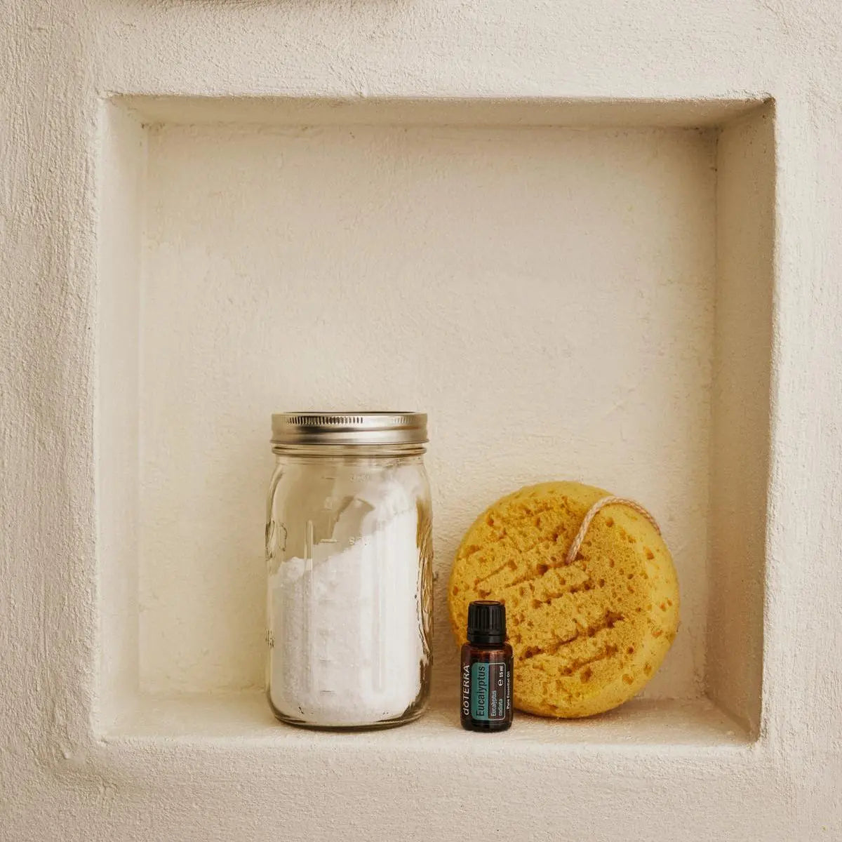 Aceite esencial de Eucalipto doTERRA 15ml en nicho de ducha de yeso blanco junto a bote de cristal con sal de bano y esponja marina natural