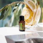 Aceite esencial de eucalipto limón dōTERRA 15ml en mesa blanca reflectante con jarra de vidrio llena de rodajas de limón y rama de eucalipto, luz natural