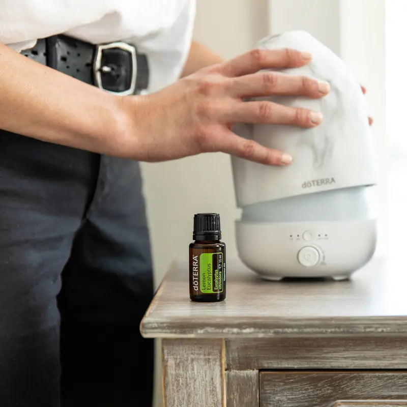 Aceite esencial de eucalipto limón dōTERRA 15ml sobre mesa de madera envejecida junto a difusor dōTERRA blanco grande con mano ajustando la tapa