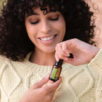 Mujer joven de pelo rizado sonriendo mientras sostiene frasco de aceite esencial de eucalipto limón dōTERRA 15ml con ambas manos, vistiendo jersey trenzado crema