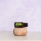 Aceite esencial de eucalipto limón dōTERRA 15ml apoyado en piedra de arcilla redondeada sobre superficie blanca, fondo pared texturizada lavanda, composición minimalista