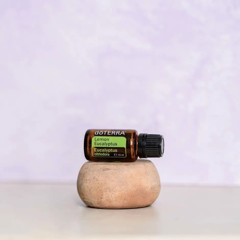 Aceite esencial de eucalipto limón dōTERRA 15ml apoyado en piedra de arcilla redondeada sobre superficie blanca, fondo pared texturizada lavanda, composición minimalista