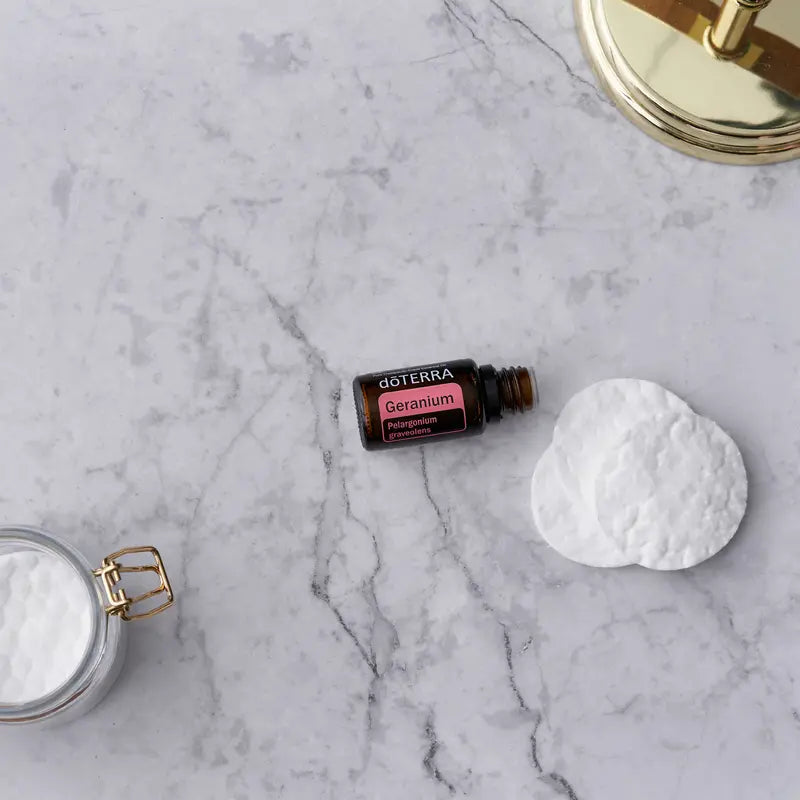 Aceite esencial de geranio dōTERRA 15ml en mármol blanco junto a algodones redondos, tarro de vidrio con tapa dorada y espejo dorado