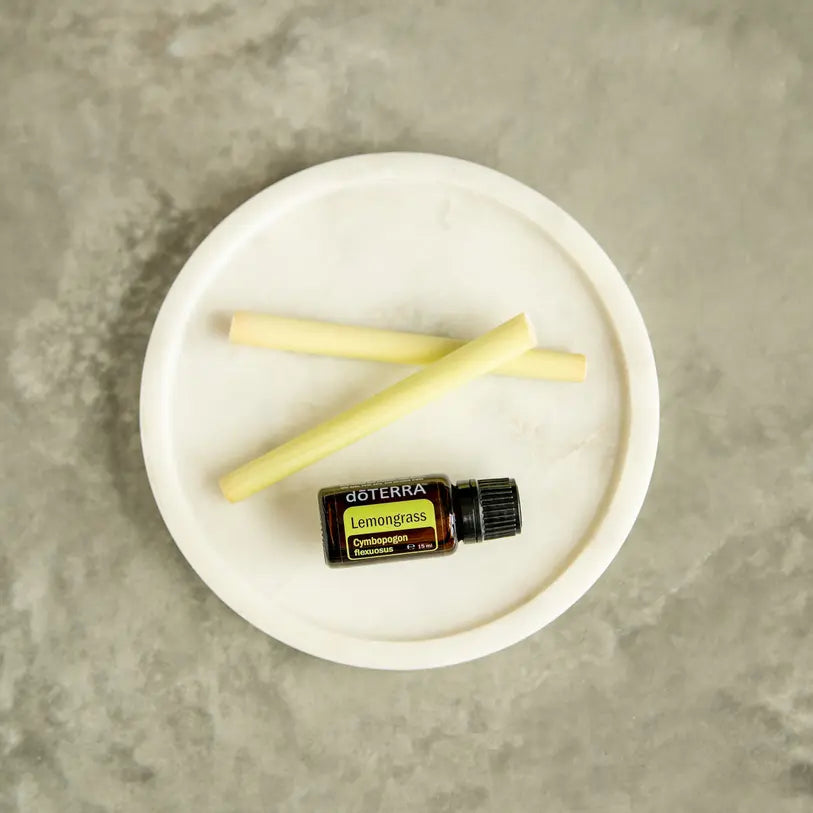 Aceite esencial de Lemongrass doTERRA 15ml sobre plato de marmol blanco junto a dos tallos frescos de hierba limon pelados