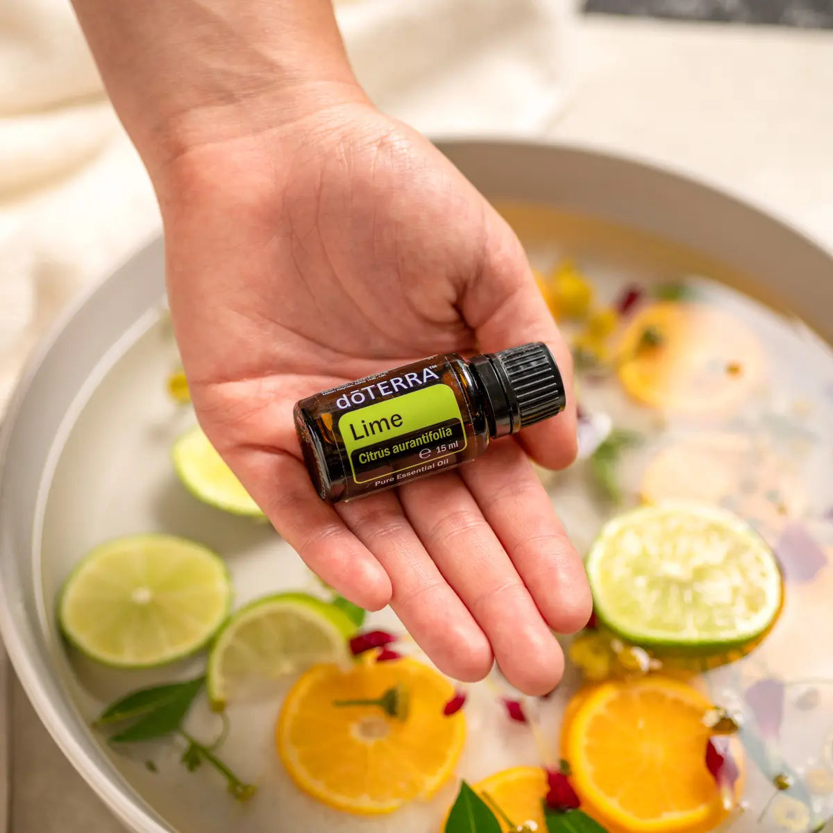 Aceite Esencial Lima dōTERRA 15ml con mano sujetando cuenco de agua con cítricos y flores