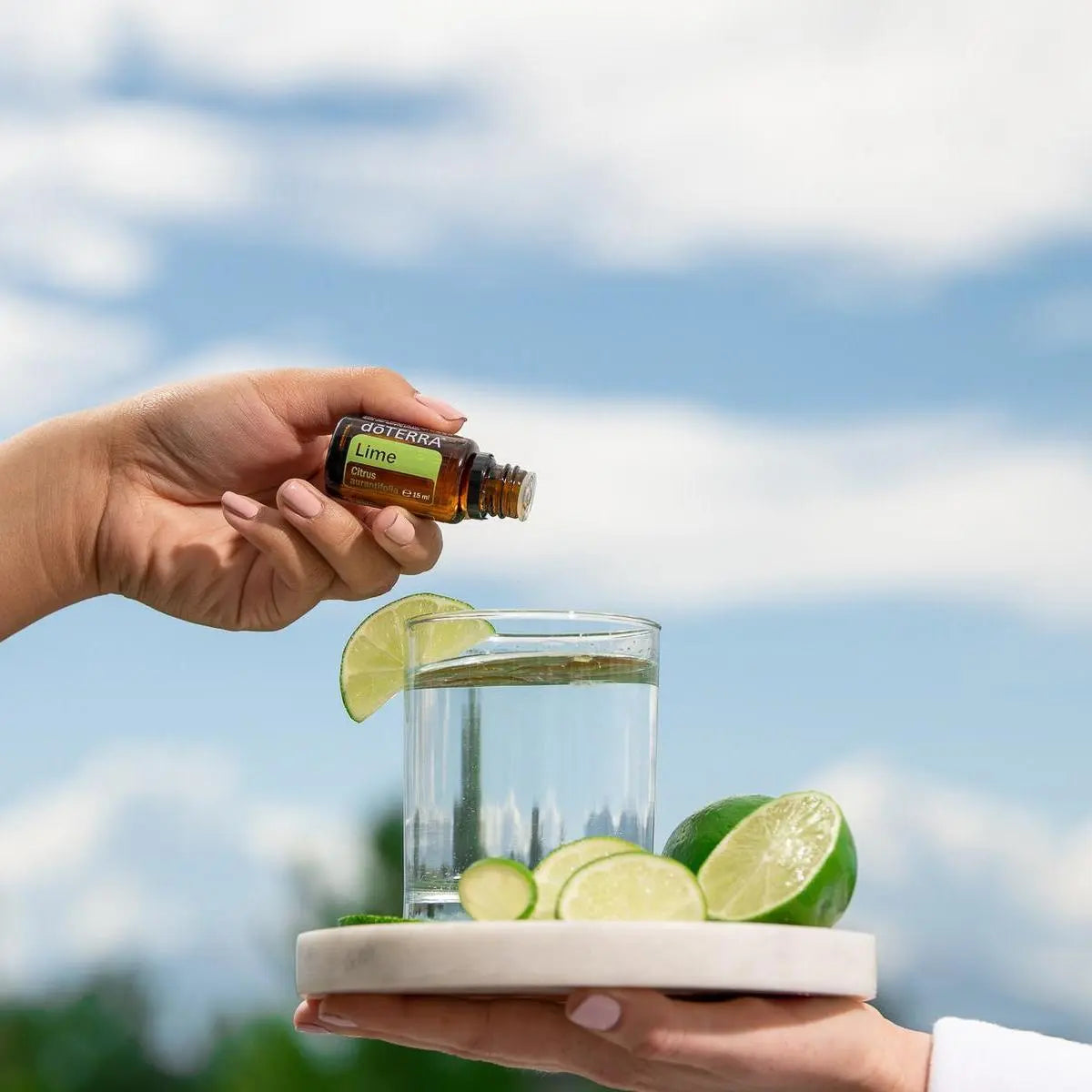 Aceite Esencial Lima dōTERRA 15ml con mano sosteniendo gota sobre vaso de agua con lima al cielo azul