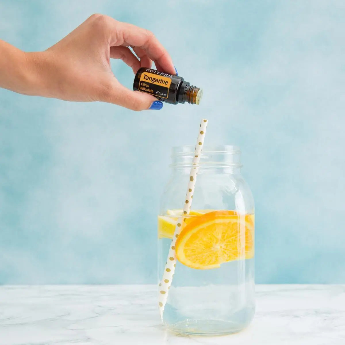 Aceite Esencial Mandarina dōTERRA 15ml con mano sosteniendo gota sobre tarro de cristal con rodajas de naranja y paja
