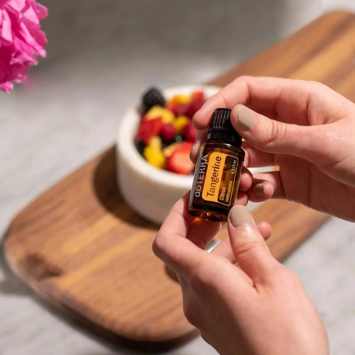 Aceite Esencial Mandarina dōTERRA 15ml con manos sosteniendo frasco en cuenco de frutas variadas sobre tabla de madera