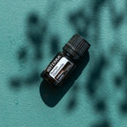 Aceite esencial de Manzanilla Romana dōTERRA 5ml sobre fondo teal con sombras dramáticas de hojas proyectadas por luz solar directa