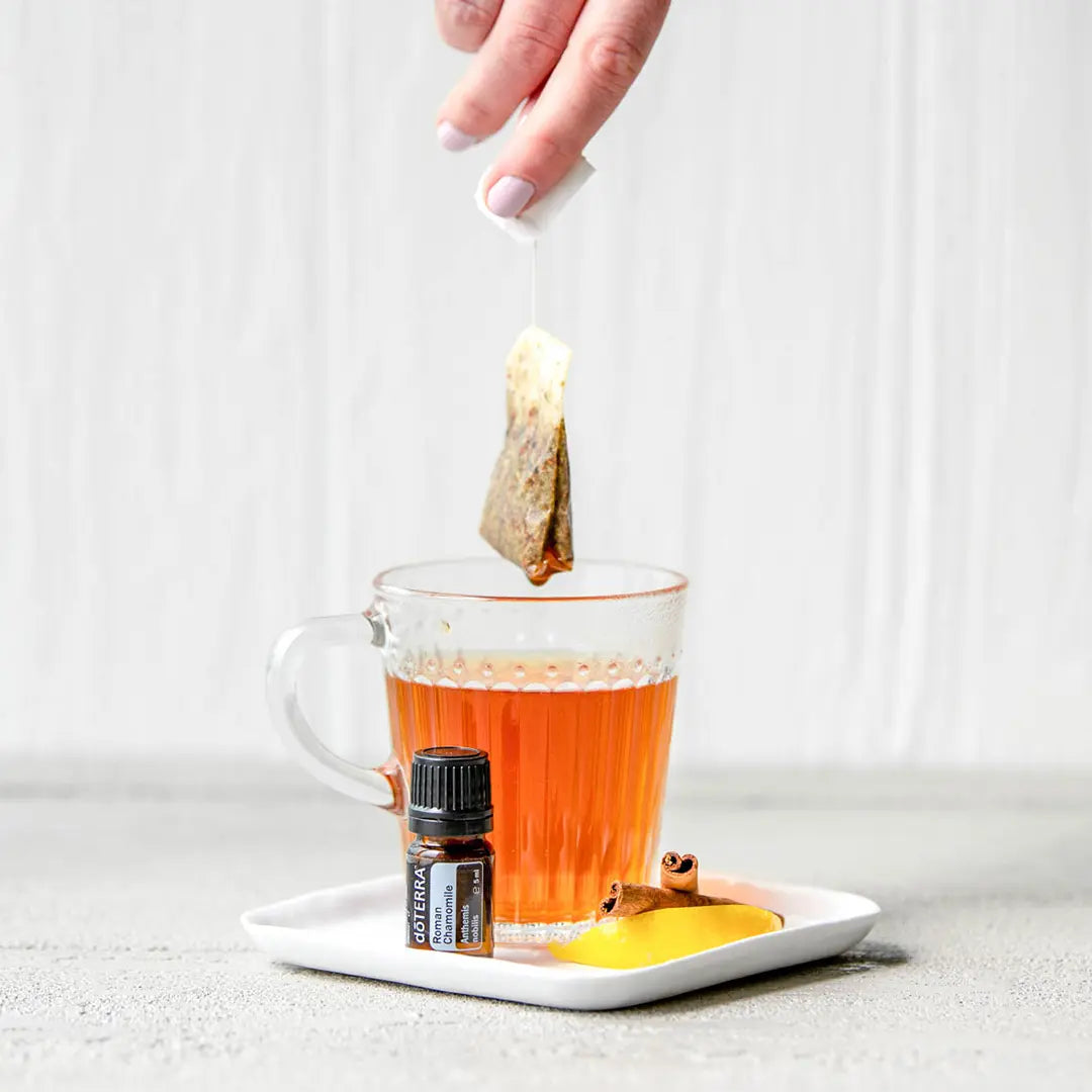 Aceite esencial de Manzanilla Romana dōTERRA 5ml junto a taza de vidrio con té e infusión, canela y platillo blanco, fondo neutro