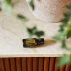 Aceite esencial de Mejorana dōTERRA 15ml horizontal sobre encimera de mármol beige con maceta de cerámica texturizada y hojas en bokeh