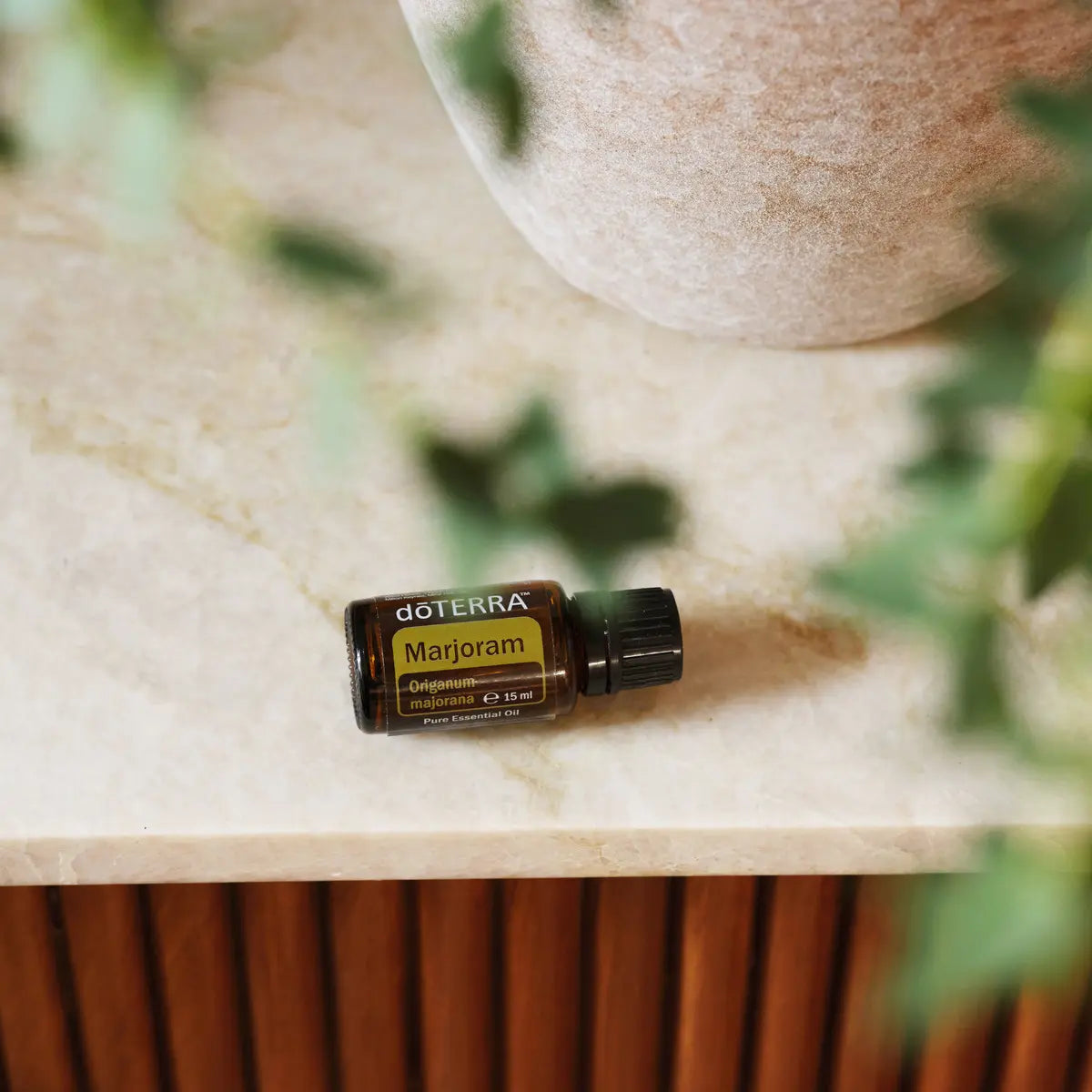 Aceite esencial de Mejorana dōTERRA 15ml horizontal sobre encimera de mármol beige con maceta de cerámica texturizada y hojas en bokeh