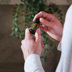 Manos con jersey blanco sosteniendo frasco de aceite esencial de Mejorana dōTERRA 15ml, planta colgante de hoja verde al fondo