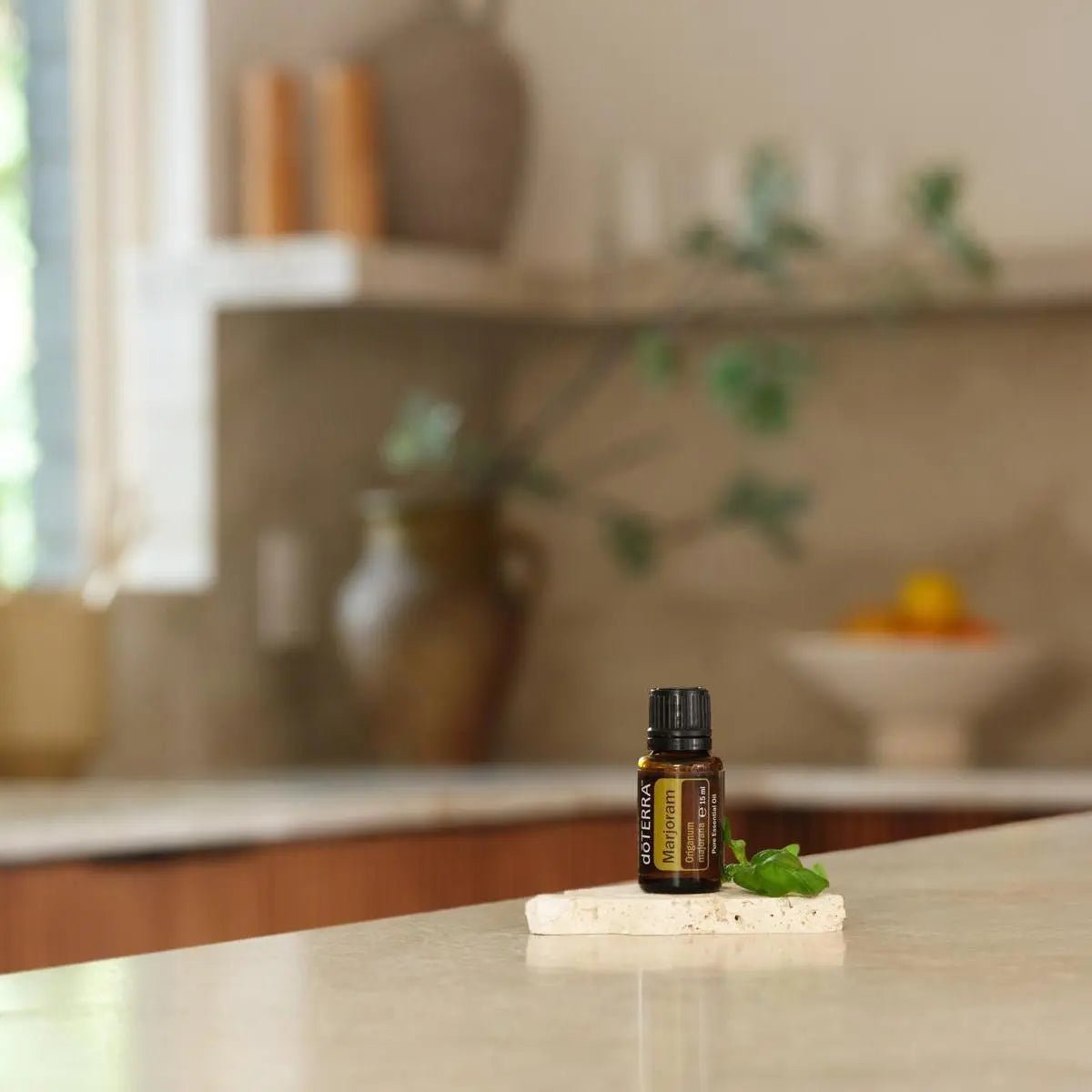 Aceite esencial de Mejorana dōTERRA 15ml sobre piedra travertino con hoja de mejorana, cocina moderna desenfocada al fondo