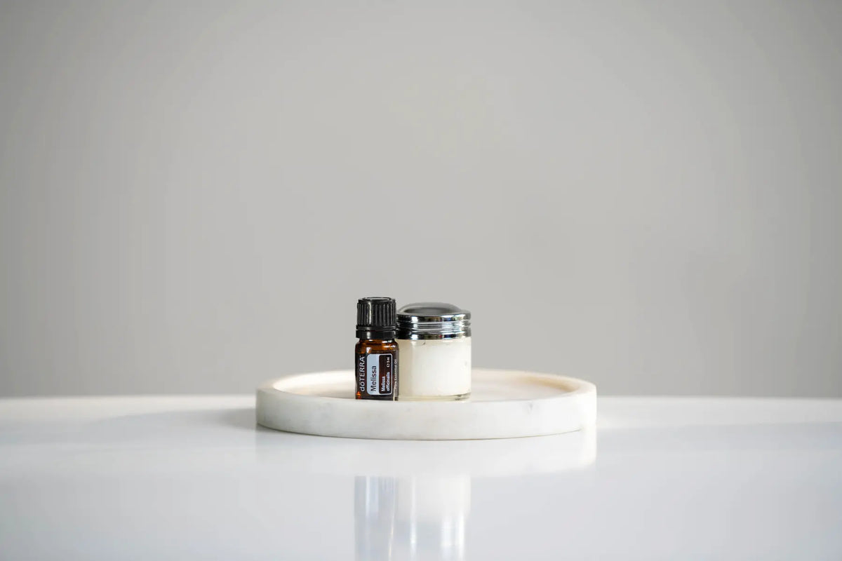 Aceite esencial de Melissa dōTERRA 5ml junto a tarro de crema sobre bandeja circular de mármol blanco, fondo gris